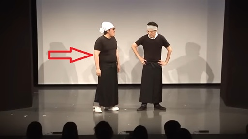  ステージ上で、白衣と黒いエプロンを着用した二人の男性が立っている。左の男性には赤い矢印が向けられている。